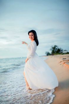 A joyful woman in a flowing white dress enjoys a serene walk along the sandy beach at sunset.