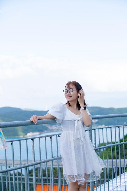 Free An asian woman in a white dress, enjoying a scenic summer view from a railing with a joyful expression. Stock Photo