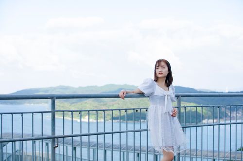 Free Asian woman in a white dress standing on a balcony with a scenic view of water and mountains. Stock Photo