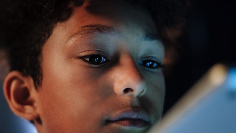 A close-up portrait of a boy with curly hair intently looking at a screen indoors.