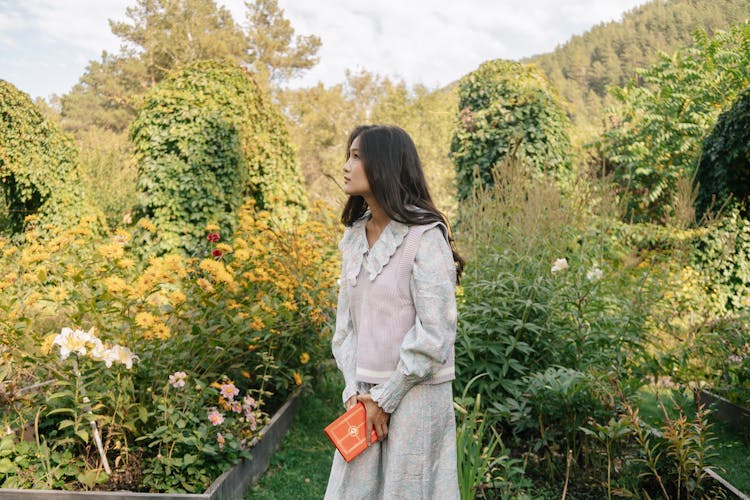 A Young Woman Holding A Book While Standing In A Garden