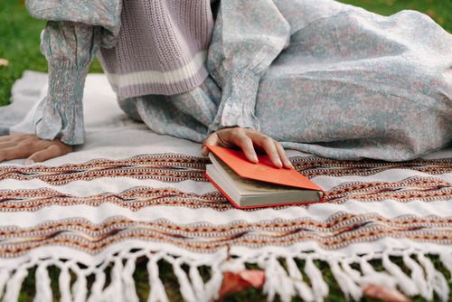Free A person in a floral dress reading a book while sitting on a patterned picnic blanket outdoors. Stock Photo