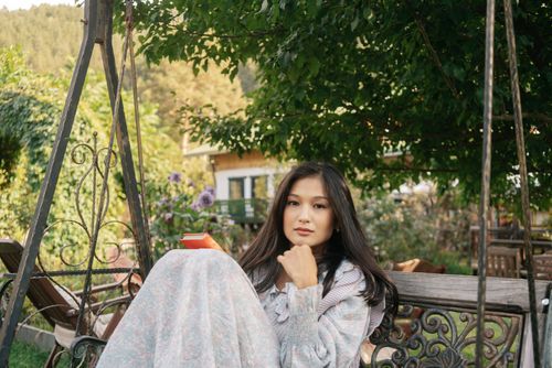 Free A young Asian woman sits on a swing in a garden, enjoying a book on a peaceful day. Stock Photo