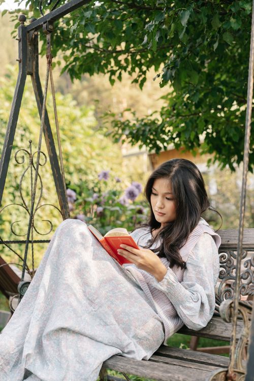 Free A serene portrait of a young woman reading a book on a garden swing surrounded by lush greenery. Stock Photo