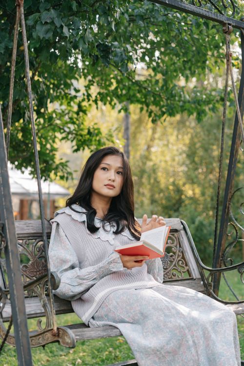 Free A serene moment with an Asian woman reading a book on a garden swing. Stock Photo