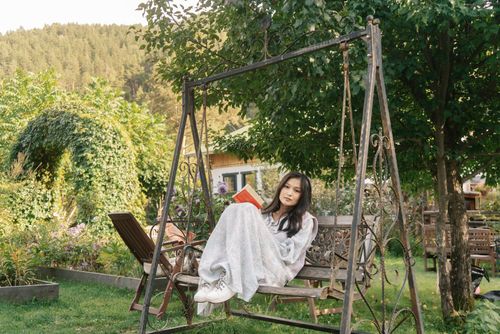 Free Young woman enjoying a book on a rustic swing in a sunlit garden setting. Stock Photo