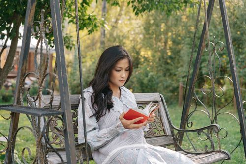 Free Asian woman reading a book while sitting on a swing in a tranquil garden setting. Stock Photo