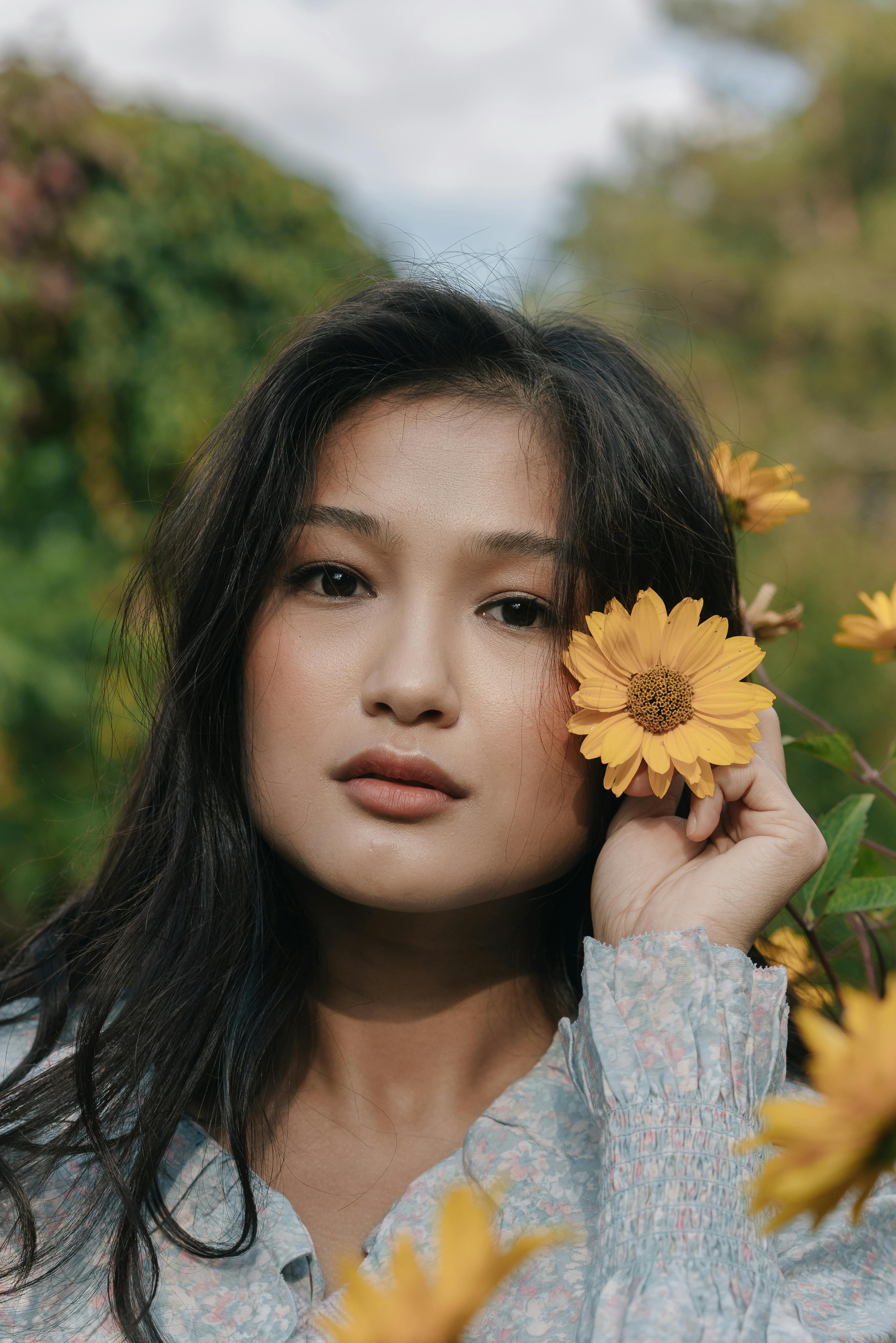 An Asian woman holding a yellow flower outdoors, showcasing natural beauty.