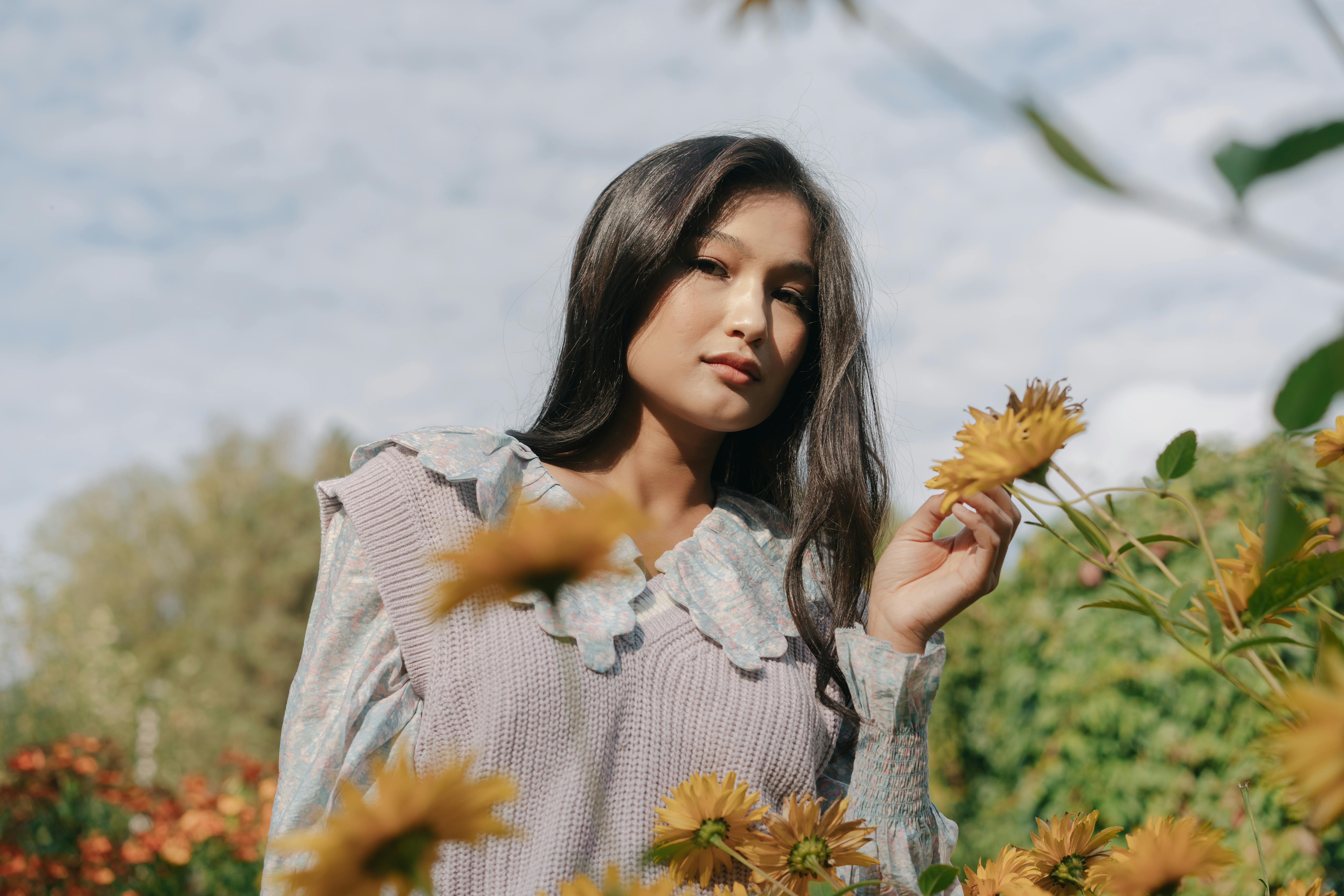 A person in a floral outfit stands among yellow flowers with a backdrop of greenery and a cloudy sky.