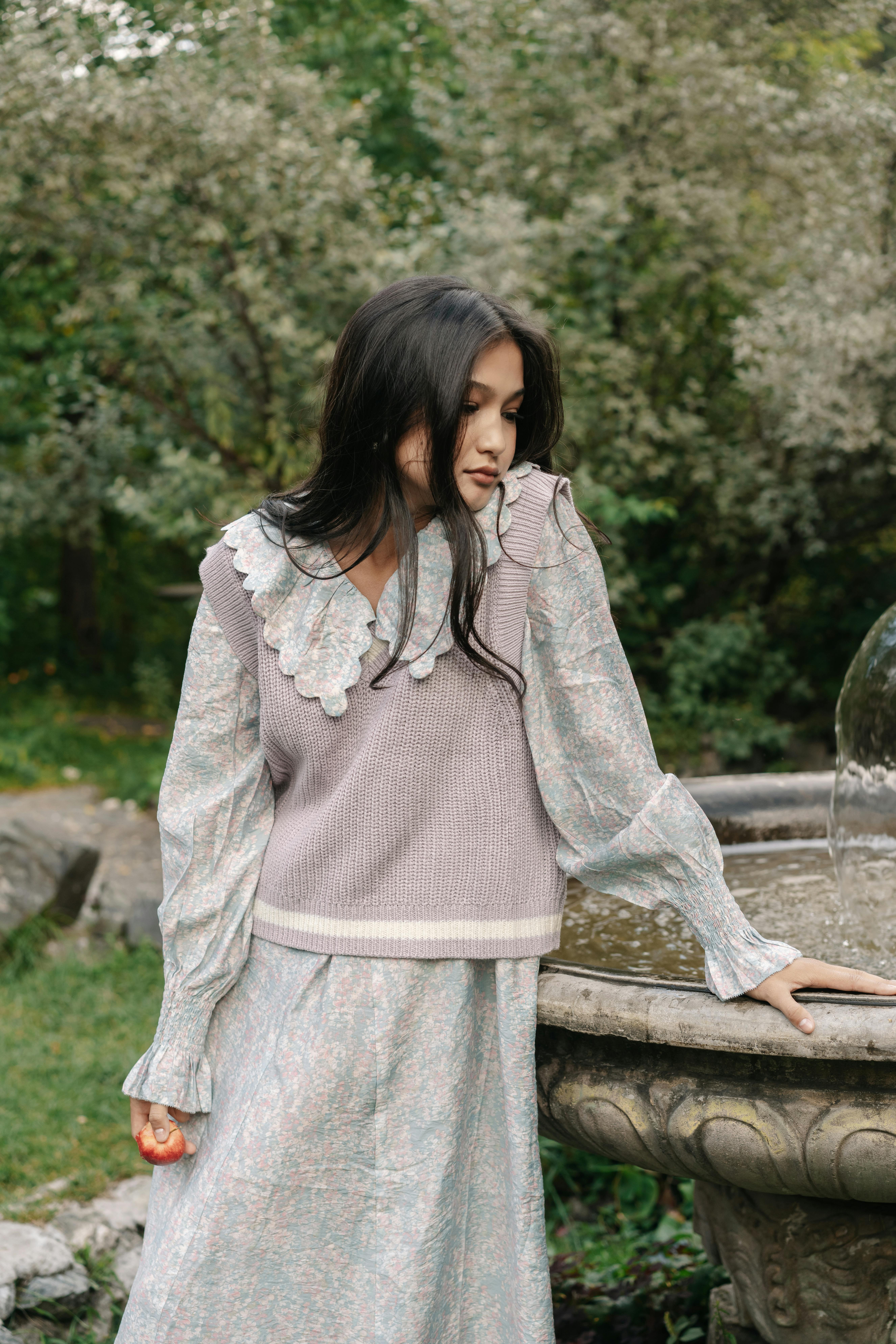 Stylish Asian woman in a vintage dress standing by a garden fountain.