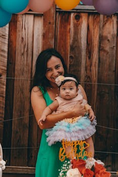 Mother holding baby at a colorful birthday party with decorations and flowers.