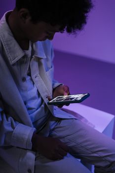Teenager focused on smartphone indoors with creative, soft purple lighting.
