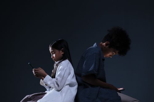 Free Two children focused on smartphones sitting back-to-back in a studio setting. Stock Photo