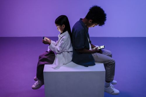 Free Two kids sitting back-to-back focused on smartphones, lit by moody lighting. Stock Photo