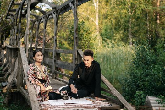 A couple in traditional kimonos performing calligraphy on a rustic wooden footbridge in a lush forest setting.