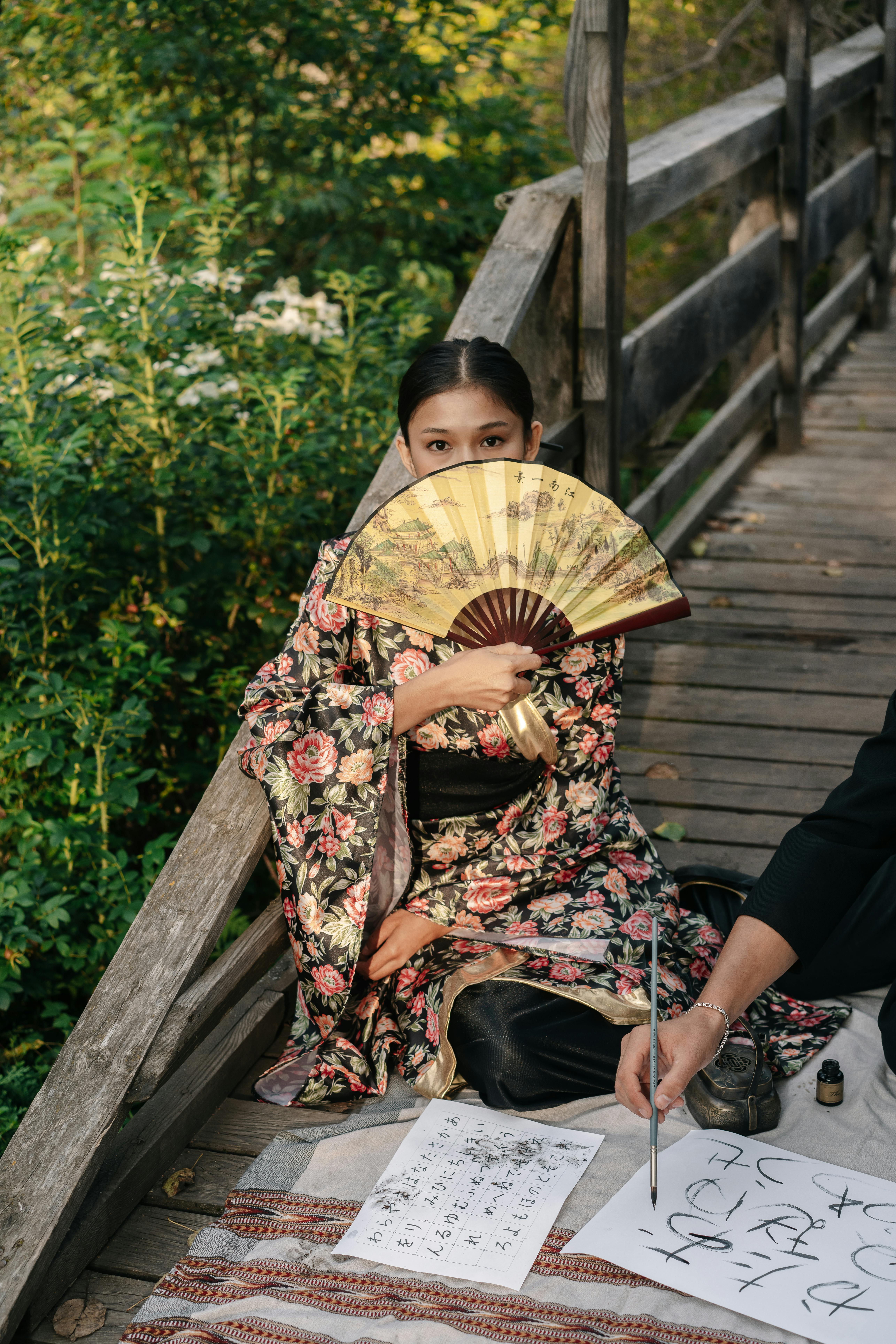 woman in a floral furisode covering her face with a fan sitting on a wooden footbridge