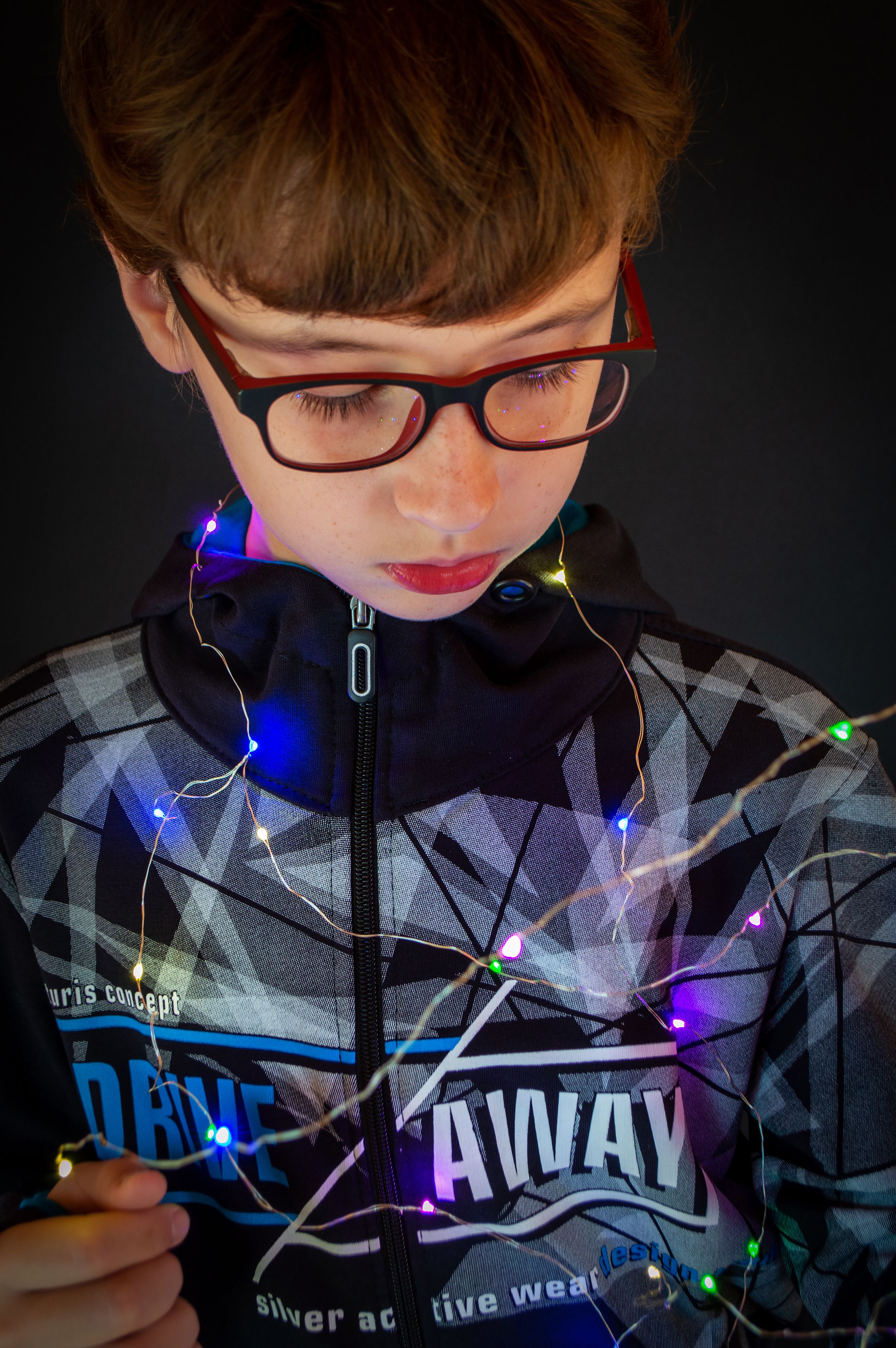 Young boy wearing eyeglasses, entangled in vibrant Christmas lights against a dark background.