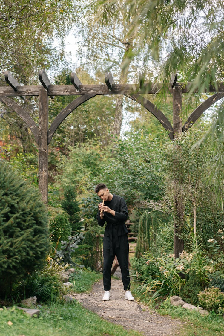 A Man In Black Clothes Standing In A Garden While Playing The Flute