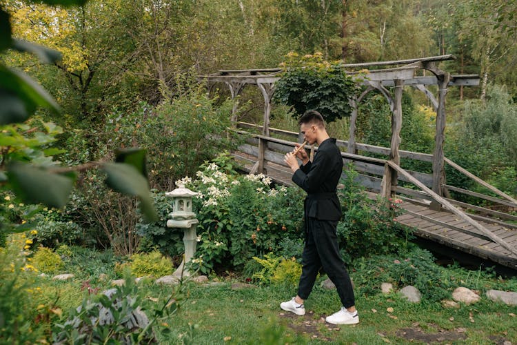 A Man In Black Clothes Walking In A Garden While Playing The Flute