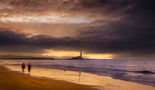 Free Couple walking on a beach with dramatic sunset and lighthouse in view. Stock Photo