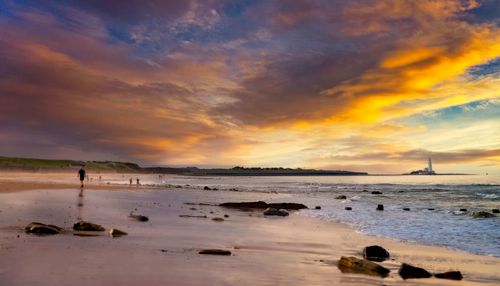 Free A breathtaking sunset over a tranquil beach with a dramatic sky and distant lighthouse. Stock Photo