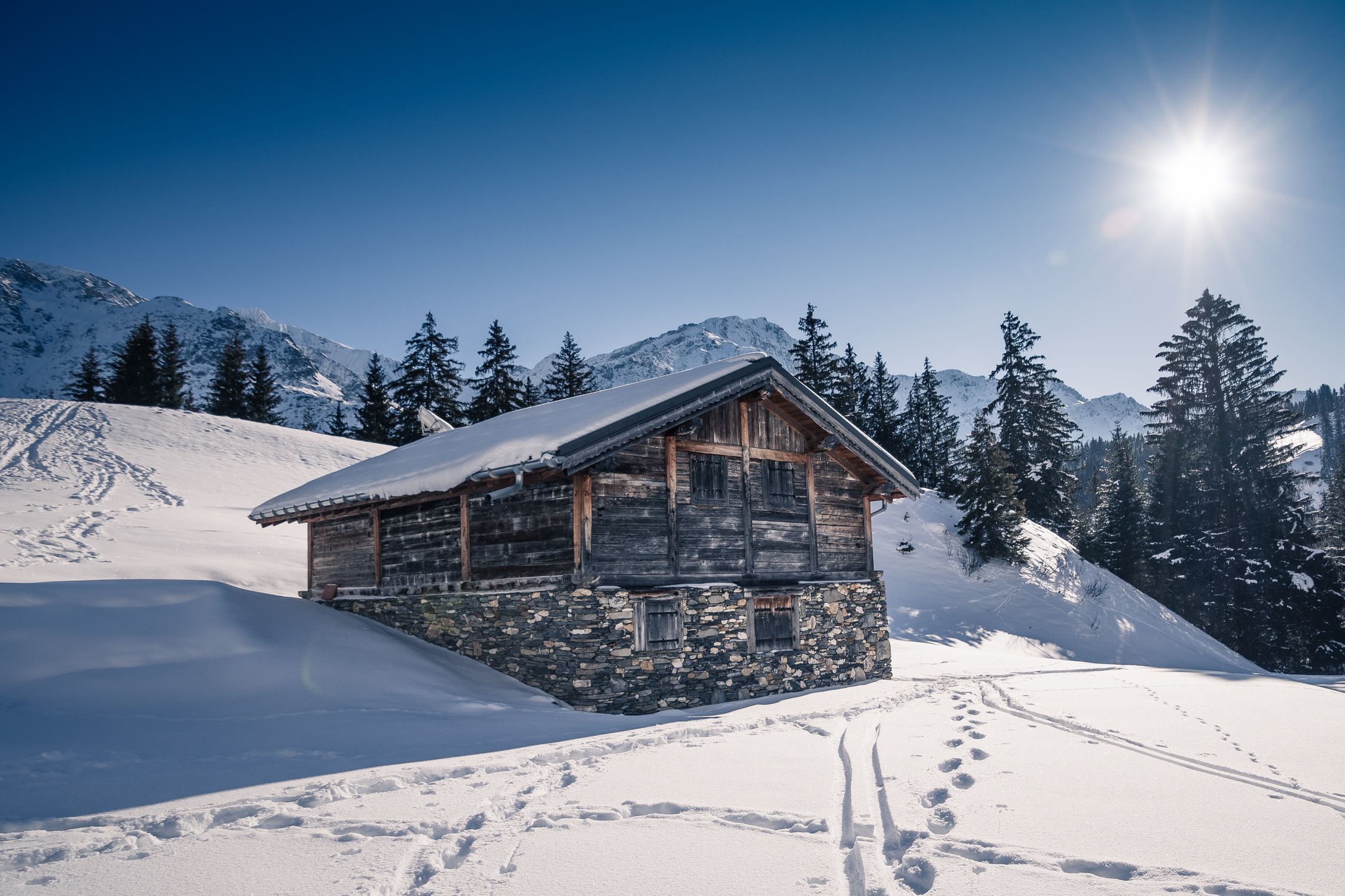 Charming rustic chalet in snowy French Alps with bright sun and mountain backdrop.