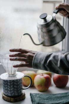 A cozy indoor scene of hand pouring hot water for coffee by a window with apples.