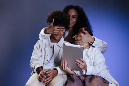A mother and her children sharing a playful moment with a tablet against a blue background.