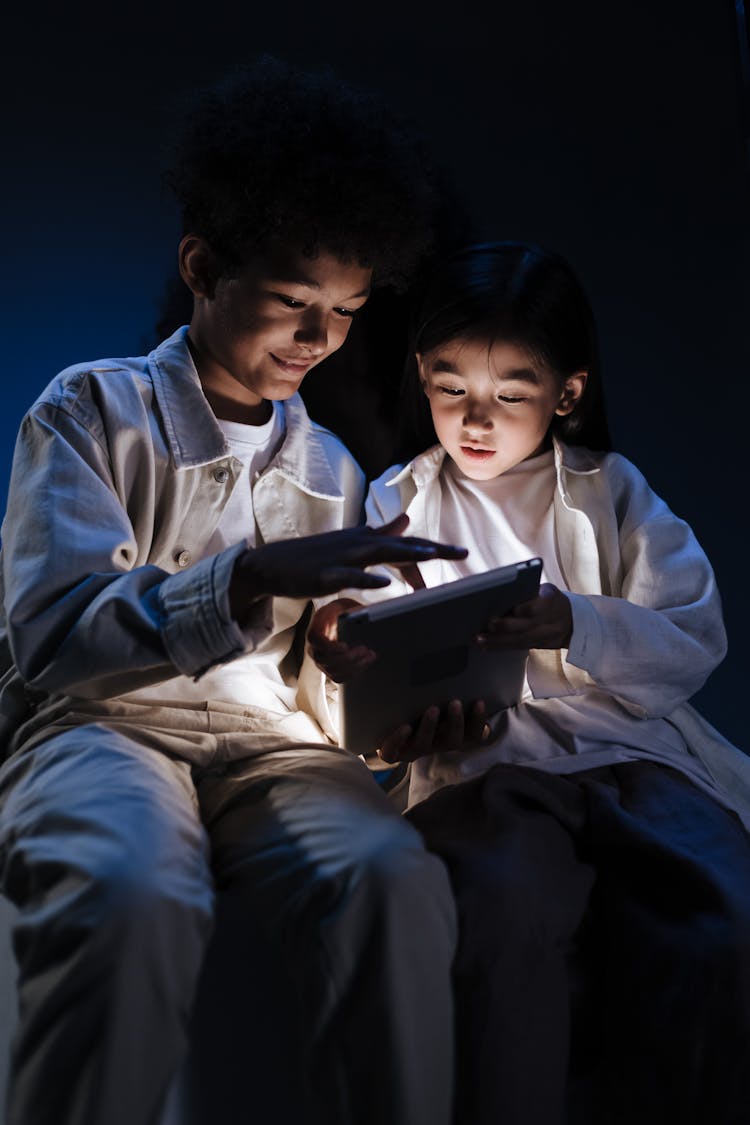 Two Kids Sitting In Dark Room And Looking With Interest To Internet On Digital Pad