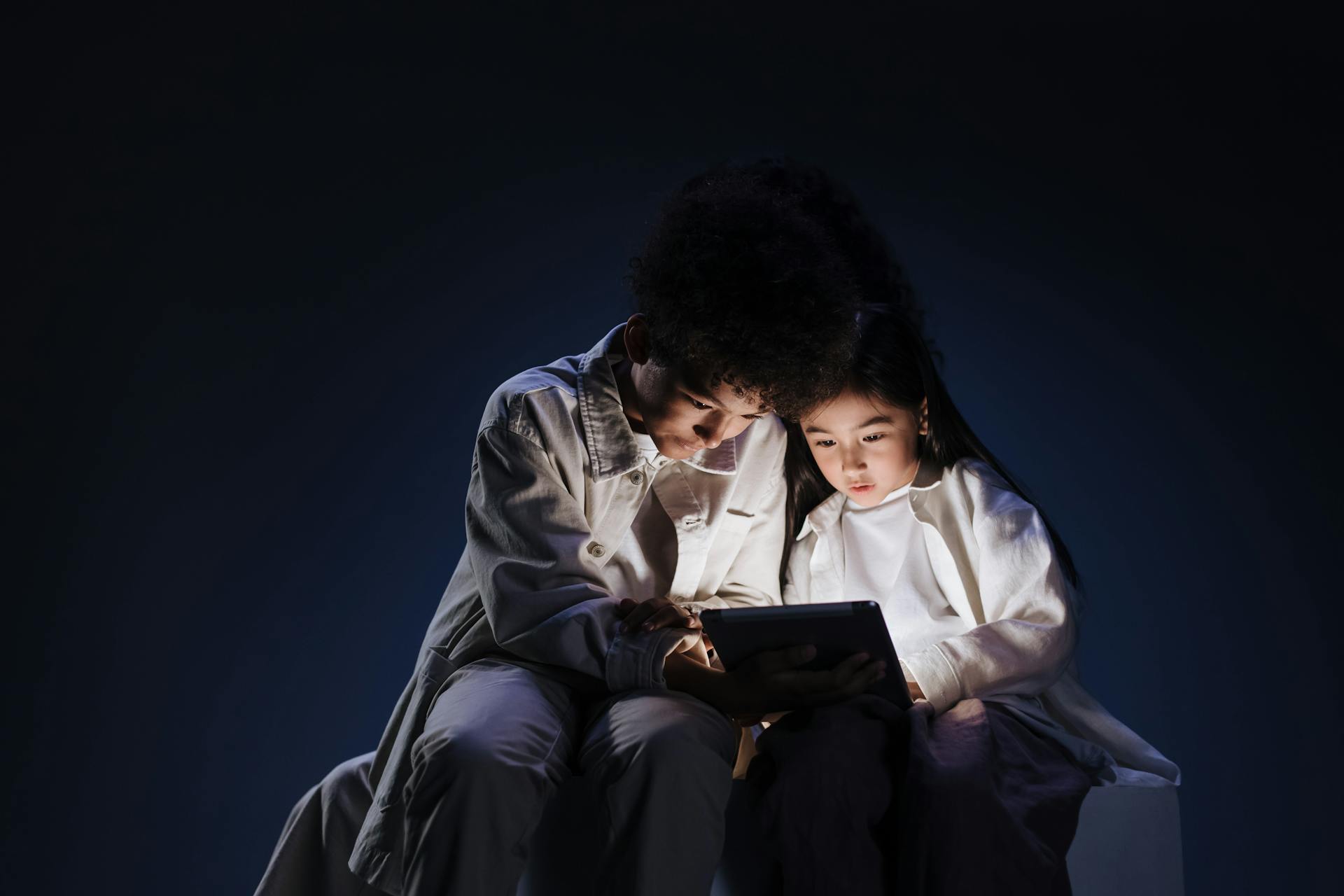 Teenage children sitting together in dark over bright screen of digital pad
