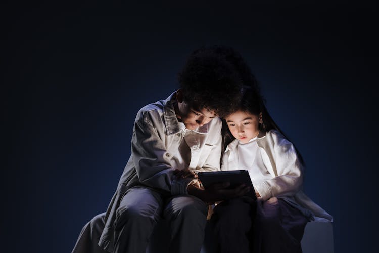 Teenage Children Sitting Together In Dark Over Bright Screen Of Digital Pad