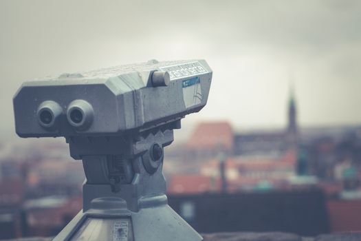 An outdoor telescope overlooking a blurred urban cityscape under a cloudy sky.