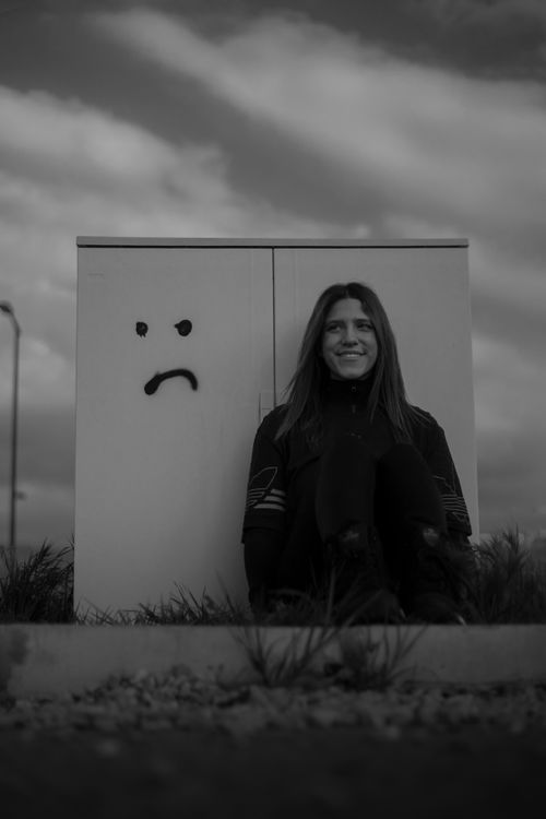 Free Black and white image of a woman sitting beside a box with a sad face graffiti. Stock Photo