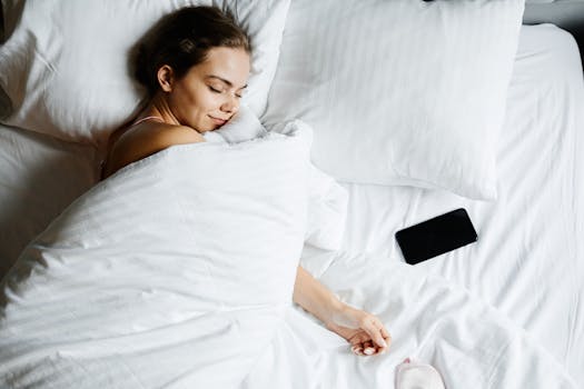 A young woman sleeps comfortably in bed with a smartphone beside her, wrapped in white sheets.