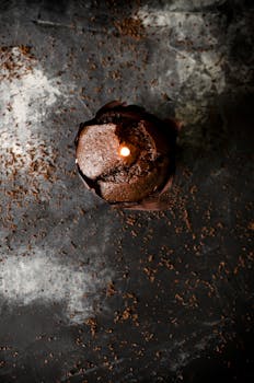 An artistic overhead shot of a chocolate muffin with a lit candle on a dark textured surface.