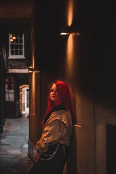 A woman with red braided hair leans against a dimly lit alleyway wall at night.