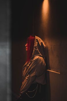 Artistic and moody portrait of a red-haired woman in a hoodie, standing against a wall in low light.