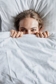 A cheerful young woman peeks from under a blanket, embracing a cozy morning in bed.