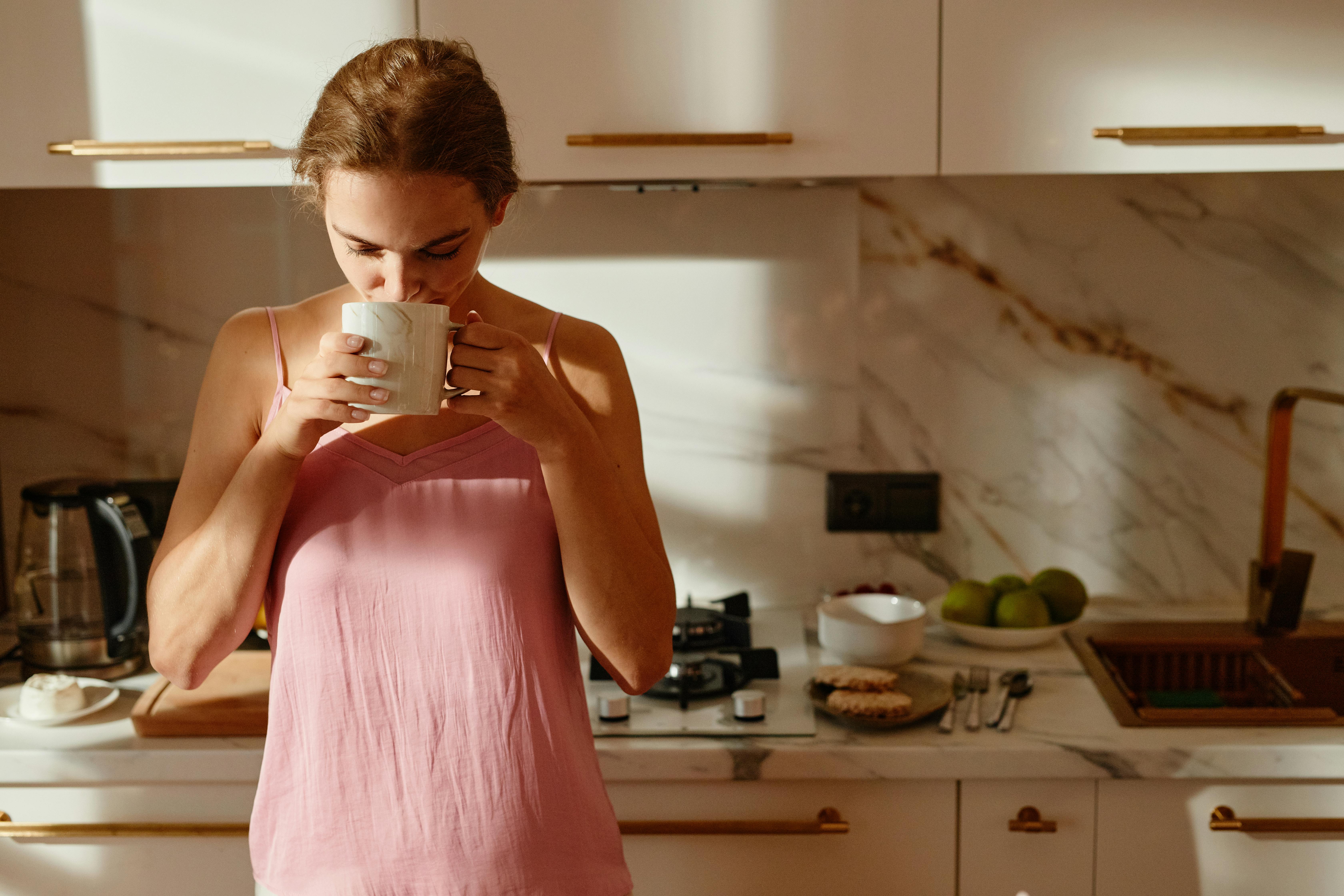 A woman enjoys a hot beverage in a sunlit kitchen, creating a tranquil morning scene.