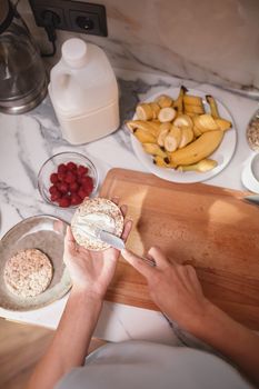 A person spreads cream on rice cakes with fresh fruit and milk on a counter, indoors.
