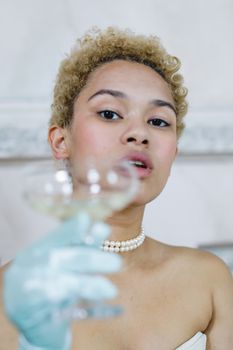 A stylish woman holding a champagne flute, wearing a pearl necklace and gloves, in an elegant setting.