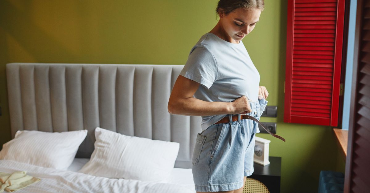 Woman adjusting her belt in a cozy bedroom, dressed in casual denim shorts and a blue shirt.