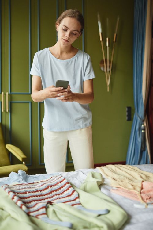 Free Young woman using smartphone to organize clothes on bed in a stylish room. Stock Photo