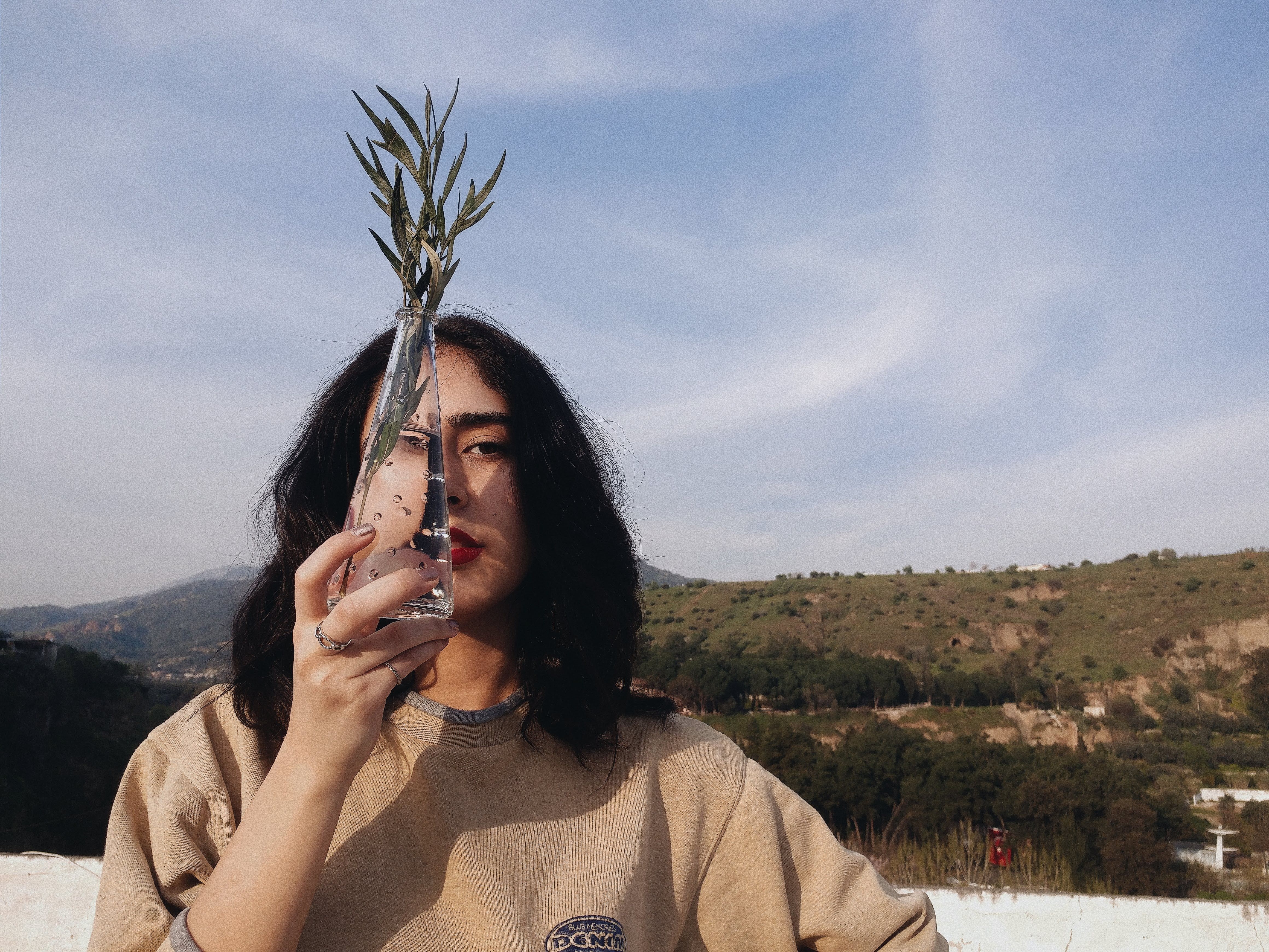 A woman stands outdoors holding a glass vase with a sprig against a blue sky backdrop.