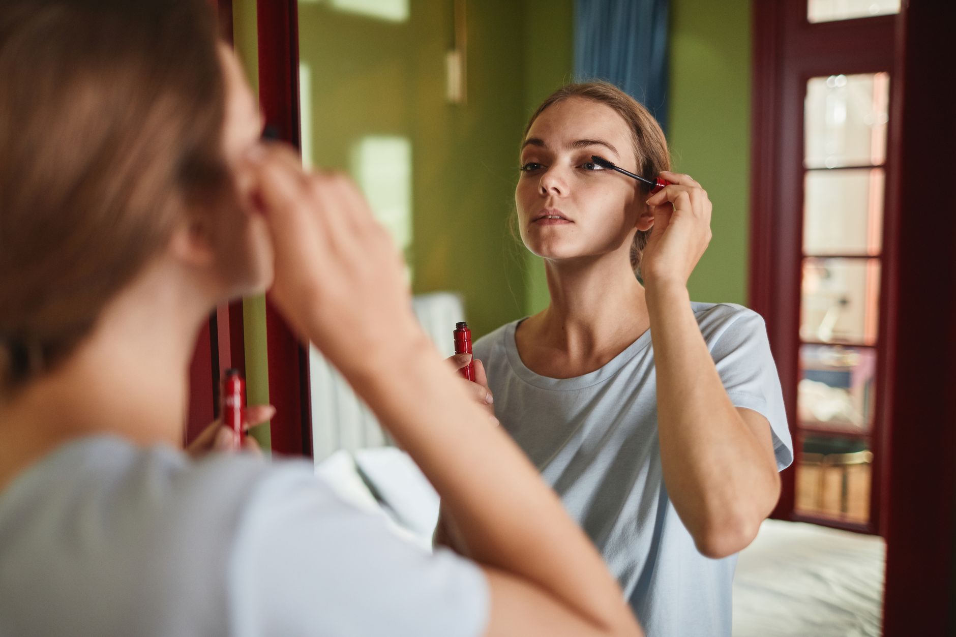 Adult woman applying mascara in front of a mirror indoors, focusing on beauty and makeup routine.