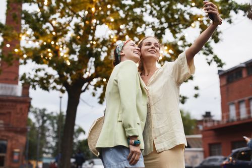 Free Two young women taking a selfie outdoors with a festive tree background in an urban setting. Stock Photo
