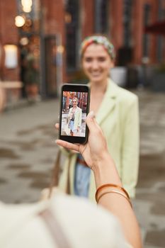 A woman in stylish attire is photographed with a smartphone outdoors on an urban street.