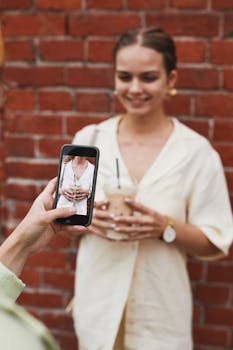 A woman holding a drink poses for a smartphone photo against a brick wall.