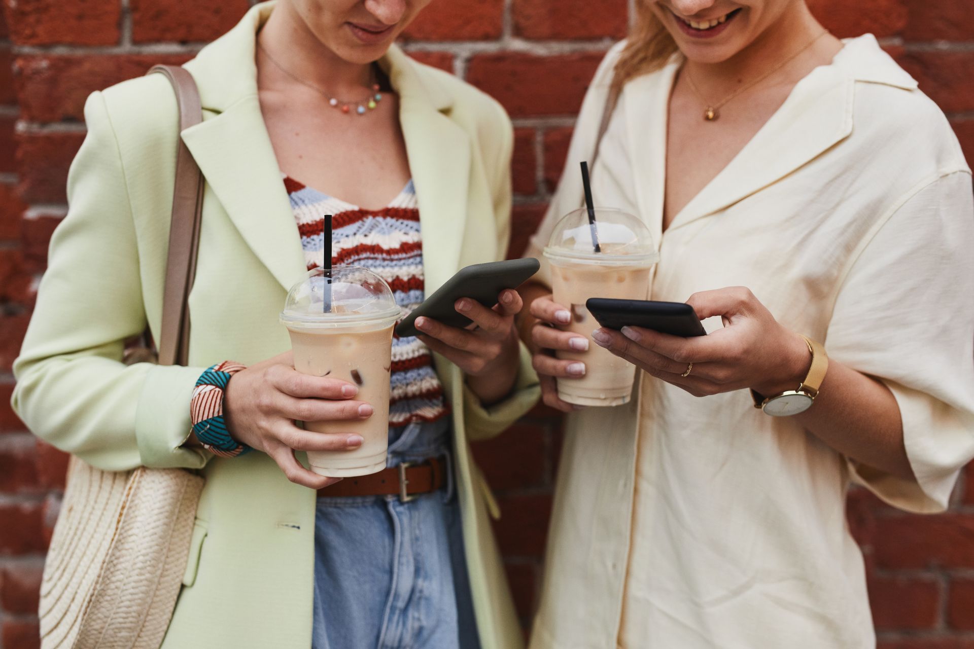 Two women enjoying iced coffee while browsing smartphones against a red brick wall.