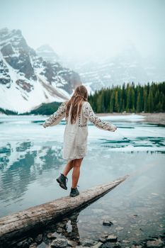A woman walks on a log by Moraine Lake, surrounded by mountains and snow in Alberta, Canada.